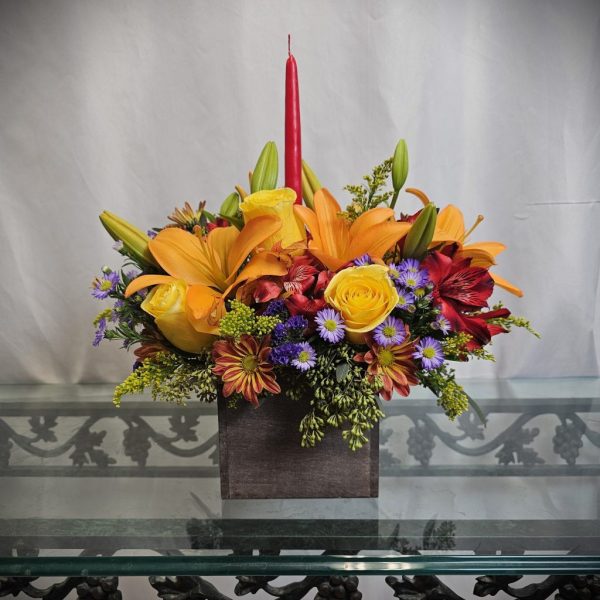 A floral arrangement with orange lilies, yellow roses, red alstroemeria, and purple asters, topped with a red candle, in a square wooden vase on a glass table.