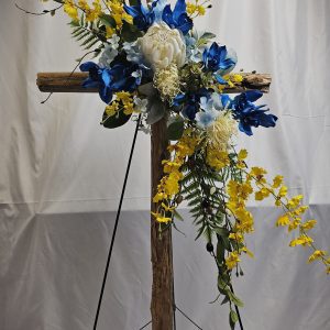 A wooden cross adorned with blue, white, and yellow flowers stands on a black easel against a white background.