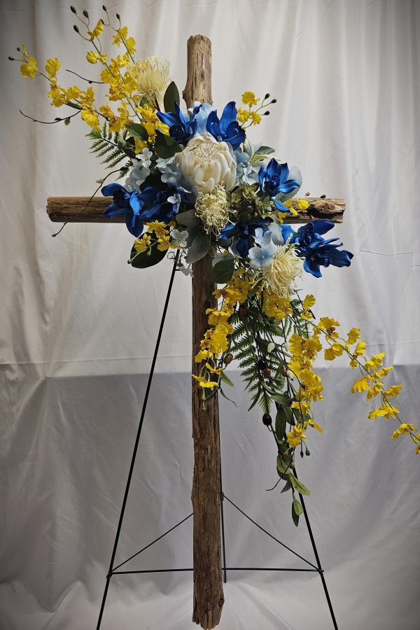 A wooden cross adorned with blue, white, and yellow flowers stands on a black easel against a white background.