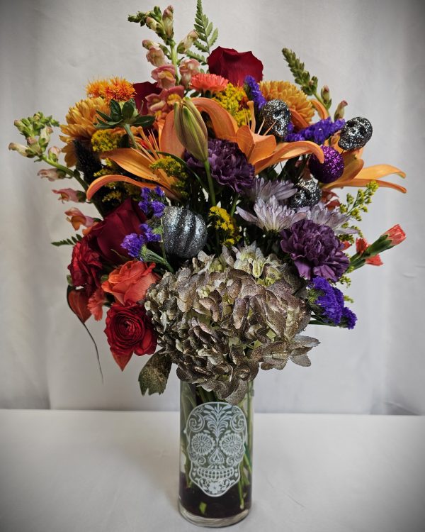 Introducing "Halloween in Bloom," a vibrant floral arrangement housed in a decorative vase adorned with a sugar skull design. This captivating bouquet showcases lilies, roses, and an assortment of flowers in stunning shades of orange, red, purple, and green.