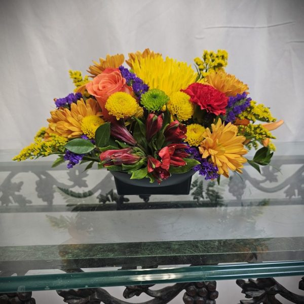 A floral arrangement featuring a variety of flowers in yellow, orange, red, purple, and green, placed on a glass table with a decorative black metal base.