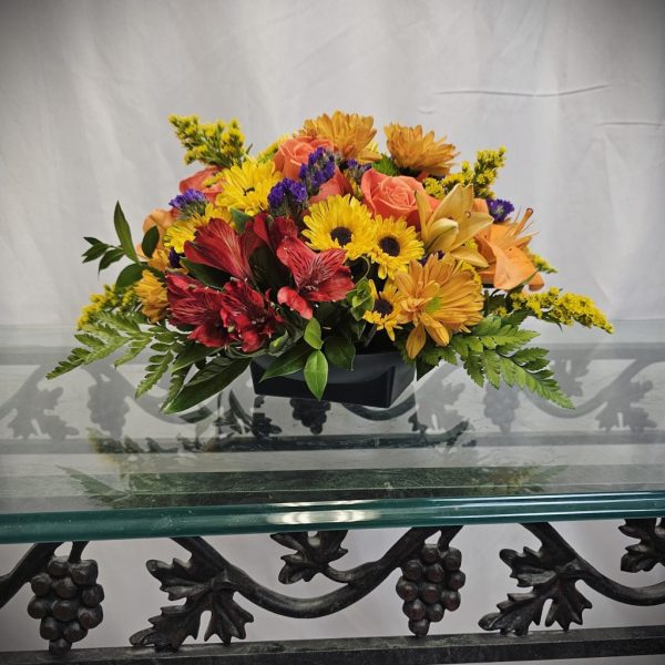 A rectangular floral arrangement with yellow, red, orange, and purple flowers on a glass table with an ornate iron base.