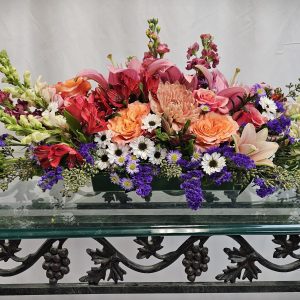 A floral arrangement featuring pink lilies, orange roses, purple statice, and white daisies, set on a glass table with a decorative wrought iron base.