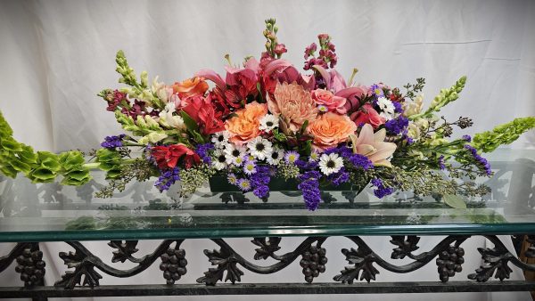 A floral arrangement featuring pink lilies, orange roses, purple statice, and white daisies, set on a glass table with a decorative wrought iron base.