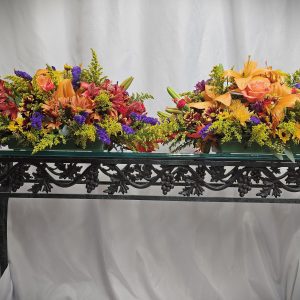 Two vibrant centerpieces from the Fall or Thanksgiving Collection adorn a decorative black metal table, showcasing orange roses, lilies, and an array of blooms in red, purple, and yellow hues against a white curtain backdrop.