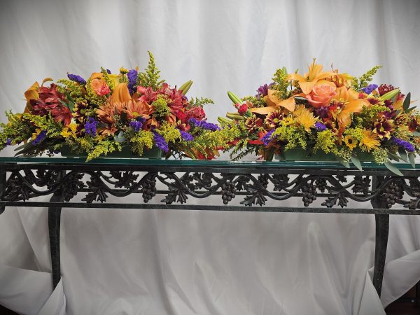 Two vibrant centerpieces from the Fall or Thanksgiving Collection adorn a decorative black metal table, showcasing orange roses, lilies, and an array of blooms in red, purple, and yellow hues against a white curtain backdrop.