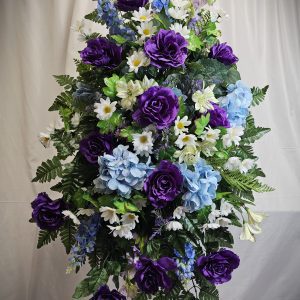 A Fresh or Silk Flower Easel featuring purple roses, blue hydrangeas, white daisies, and green foliage on a stand against a white background.