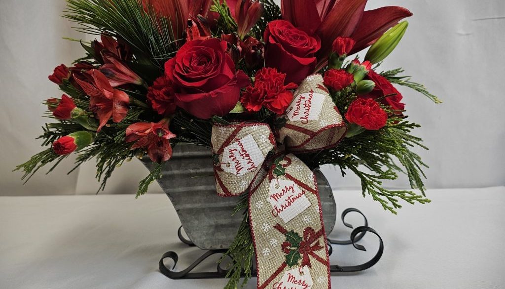 Festive floral arrangement with red lilies, roses, and carnations in a silver sleigh vase, accented with evergreen branches and a "Merry Christmas" ribbon.