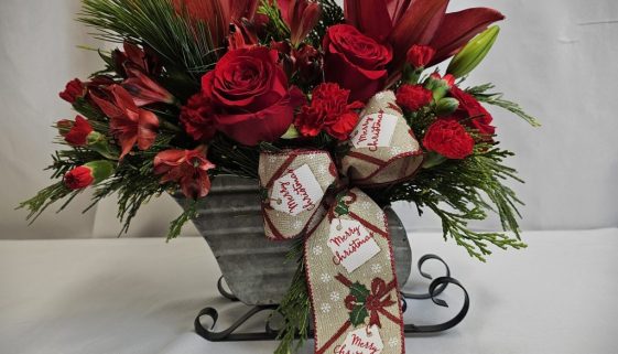 Festive floral arrangement with red lilies, roses, and carnations in a silver sleigh vase, accented with evergreen branches and a "Merry Christmas" ribbon.