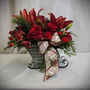 Festive floral arrangement with red lilies, roses, and carnations in a silver sleigh vase, accented with evergreen branches and a "Merry Christmas" ribbon.