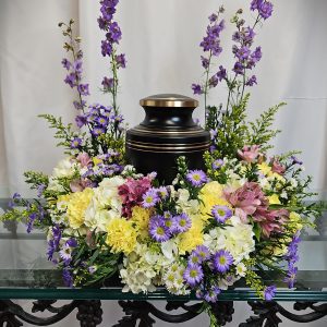 Urn surrounded by a floral arrangement with purple, yellow, and white flowers on a glass-topped table.