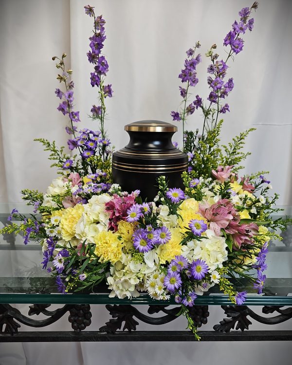 Urn surrounded by a floral arrangement with purple, yellow, and white flowers on a glass-topped table.