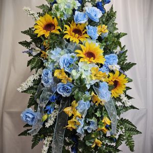 A colorful floral arrangement with sunflowers, blue roses, and various greenery set against a plain background.