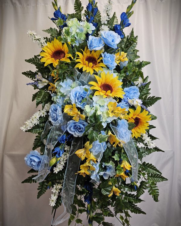 A colorful floral arrangement with sunflowers, blue roses, and various greenery set against a plain background.