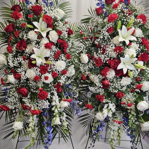 Two large "Easel Sprays" featuring red and white roses, white lilies, and blue accents are beautifully displayed against a white curtain backdrop.