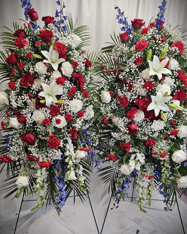 Two large "Easel Sprays" featuring red and white roses, white lilies, and blue accents are beautifully displayed against a white curtain backdrop.