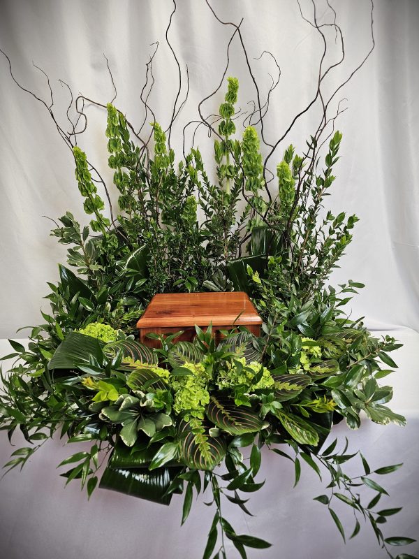 The "Wreath - Celebration of Life" features a wooden urn encircled by lush green foliage and branches against a plain white background.