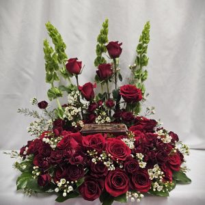 Floral arrangement with red roses, greenery, and white baby's breath surrounding a small rectangular dark brown box.