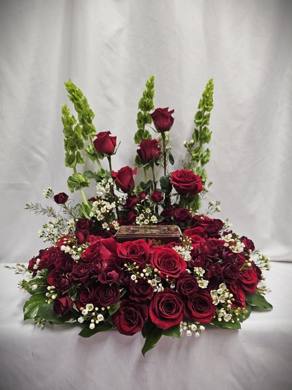 Floral arrangement with red roses, greenery, and white baby's breath surrounding a small rectangular dark brown box.